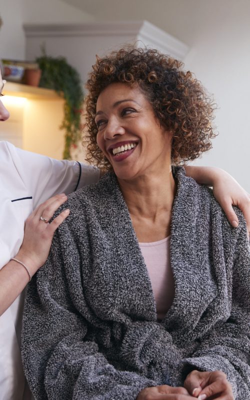 Mature Woman In Dressing Gown Talking With Female Nurse In Kitchen At Home